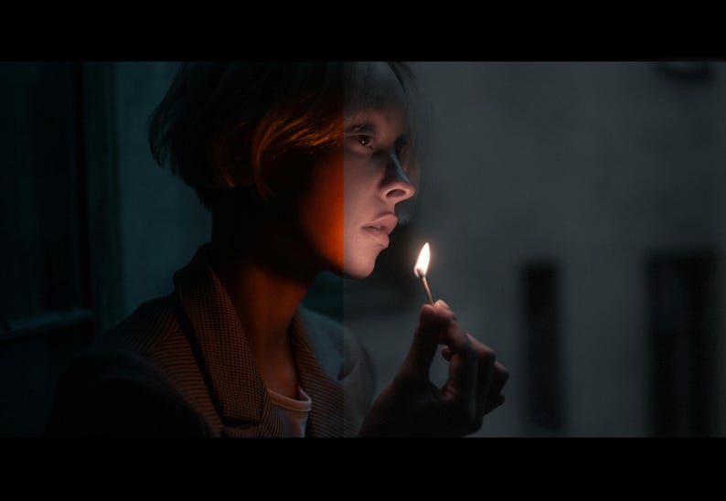 May include: A person with short, light brown hair holds a lit match in their right hand. The person is looking to the right of the frame. The person's face is partially obscured by a shadow, but the left side of their face is painted with a white and orange stripe.