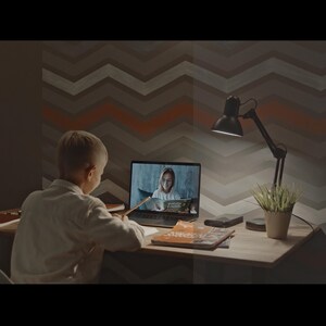 以下が含まれることがあります： 子供が机に座り、ビデオ通話を表示するラップトップの画面を見ています。デスクランプと小さな鉢植えの植物が見えます。背景は、グレーとオレンジの色合いのシェブロンパターンです。
