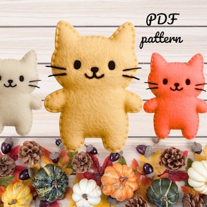 Può includere: Modello PDF per tre giocattoli di feltro a forma di gatto. I gatti sono di colori diversi: bianco, giallo e arancione. I gatti hanno occhi neri e un naso nero. Lo sfondo è in legno chiaro.