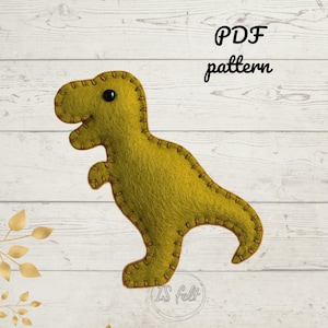 May include: A felt dinosaur in a light green color with brown stitching and a black eye. The text "PDF pattern" is written above the dinosaur. The dinosaur is on a white wooden background.