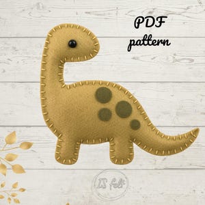 May include: A tan felt dinosaur with dark green spots and a black eye. The dinosaur is stitched with tan thread and has the words "PDF pattern" in black script. The background is a light wood grain.