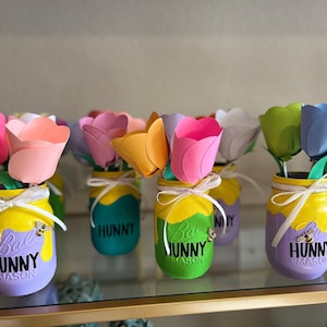 Puede incluir: Tulipanes de papel de colores en tarros Mason pintados. Los tarros están decorados en amarillo, turquesa, verde y morado, con un diseño temático de miel y la palabra "HUNNY". Cada tarro está atado con una cinta blanca.