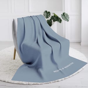 May include: A light blue blanket draped over a gray chair. The blanket has the phrase "RESTING IS PRODUCTIVE" printed in white. The chair sits on a white, round rug.