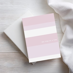 Hardcover A5 journal with striped, light pink and white design. Stripes in varying widths in three colours – light pink, lilac, and white. The encouraging words ‘One step at a time’ display in white font.