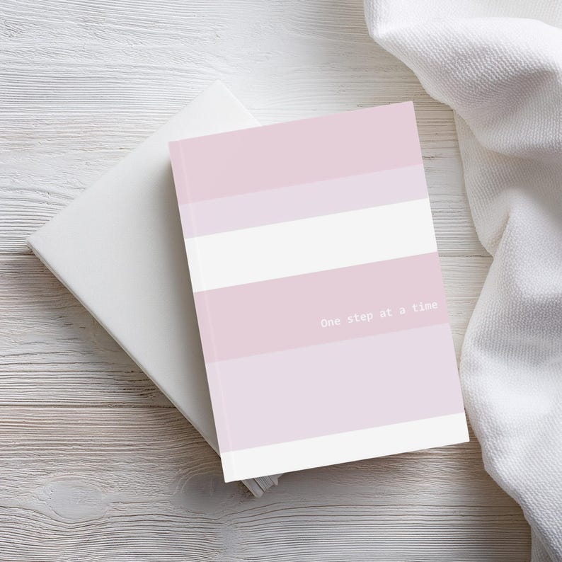 Hardcover A5 journal with striped, light pink and white design. Stripes in varying widths in three colours – light pink, lilac, and white. The encouraging words ‘One step at a time’ display in white font.