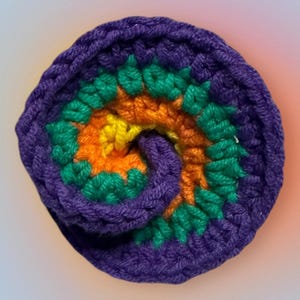 May include: A crocheted rainbow swirl coaster in shades of purple, green, orange, and yellow. The coaster is made with a thick yarn and has a textured surface.