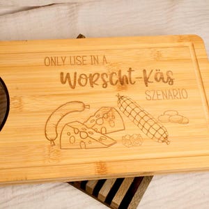 Könnte beinhalten: Ein Holz-Schneidebrett mit einem schwarzen Griff. Das Brett hat den Text "Only use in a Wurst-Käse scenario" eingraviert. Das Brett ist mit einer Zeichnung von einer Wurst, Käse und Mais verziert.