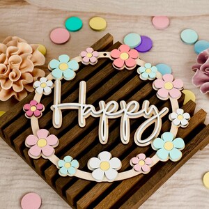 Könnte beinhalten: Ein floraler Holzkranz mit dem Wort "Happy" in der Mitte. Der Kranz ist mit pastellfarbenen Blumen in Rosa, Blau und Weiß geschmückt. Der Kranz liegt auf einer dunkelbraunen Holzbasis, mit verstreuten Konfetti.