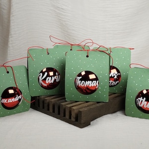 Op de afbeelding: Groene cadeaulabels met witte stippen en een rood kerstbalontwerp. Elk label heeft een naam op de ornament afgedrukt. De labels zijn vastgebonden met rood touw en staan op een houten standaard.