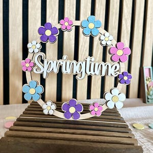 Puede incluir: Corona de primavera de madera con detalles florales coloridos en morado, rosa, azul y blanco. La palabra "Springtime" est&aacute; recortada en una fuente cursiva. La corona se exhibe sobre un soporte de madera.