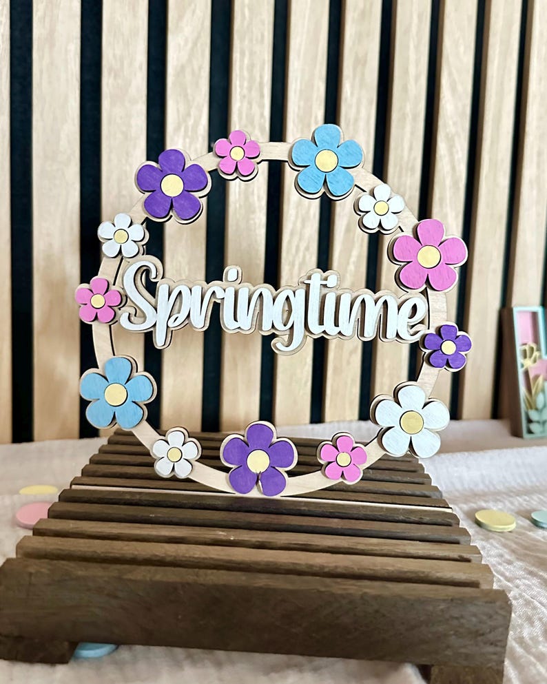 Puede incluir: Corona de primavera de madera con detalles florales coloridos en morado, rosa, azul y blanco. La palabra "Springtime" est&aacute; recortada en una fuente cursiva. La corona se exhibe sobre un soporte de madera.