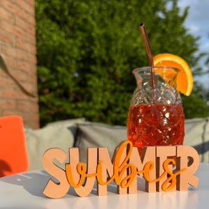 Könnte beinhalten: Ein Holzschild mit den Worten "Summer Vibes" in orangefarbenen Buchstaben. Das Schild steht auf einem weißen Tisch mit einem Glas Orangensaft und einem Strohhalm im Hintergrund.