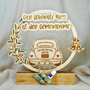 Op de afbeelding: Houten bord met een bloemmotief en een cartoonauto met de tekst "Der schönste Weg ist der gemeinsame" en "Just Married". De auto heeft een "Love" kentekenplaat en is bevestigd aan een houten standaard met hangende geld enveloppen.