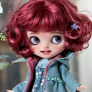 May include: A doll with short, curly magenta hair and light blue eyes. The doll has freckles, a small open mouth showing two teeth, and wears a teal corduroy hooded jacket over a floral print dress with a light blue and white polka dot collar.