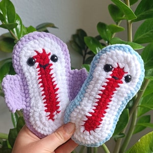 May include: Two handmade crochet ghost plushies. One is lavender, white, and red, the other is light blue, white, and red. Both have black safety eyes and a red mouth. The plushies are held in a hand.