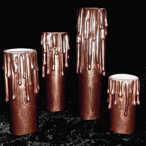 May include: Four copper-colored candles with dripping wax. The candles are different sizes and are arranged in a row on a black surface.