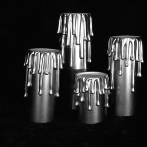 May include: A set of four cylindrical candle holders in a metallic silver finish. Each holder features a decorative design of simulated wax drips cascading from the top rim down the sides, creating a uniform, textured appearance across all pieces.