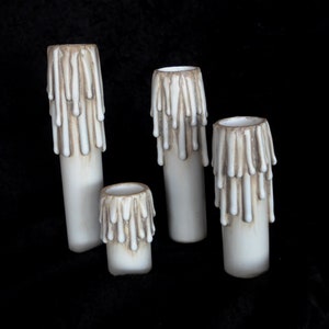 May include: Four white ceramic candle holders with a dripping wax design. The candle holders are different sizes and are arranged in a row on a black surface.
