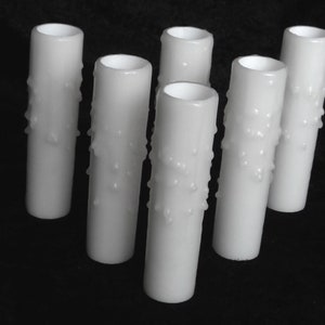 May include: Six white candle sleeves with a dripped wax design. The candle sleeves are cylindrical and have a smooth, glossy finish. They are arranged on a black surface.