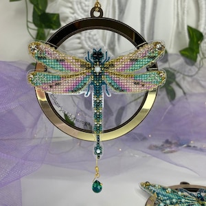 Op de afbeelding: Een decoratief libellenornament met een goudkleurige metalen ring. De libelle heeft kralen vleugels in tinten roze, blauwgroen en goud. Een groene, druppelvormige edelsteen hangt aan de onderkant. De tekst "Suzir'ya Edynoroga" staat op de zijkant.