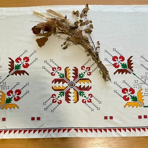 Może przedstawiać: Biały obrus lniany z haftowanymi wzorami kwiatowymi w kolorze czerwonym, żółtym, zielonym i brązowym. Obrus ma czerwoną i białą obwódkę.