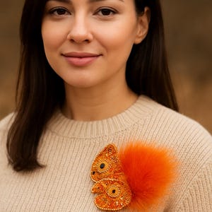 May include: A beaded squirrel brooch with an orange fluffy tail. The squirrel is made of orange and gold beads with black bead eyes. The brooch is pinned to a beige sweater.