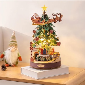 May include: A wooden Christmas tree decoration with Santa in a sleigh pulled by reindeer. The tree has lights, presents, and a train set at the base. A gnome doll and pine cone are on the left.