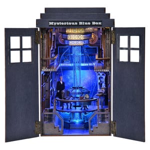 May include: A detailed wooden model of a blue box with the text "Mysterious Blue Box" on top. The interior is lit with blue and yellow lights, featuring a figure and intricate details. The doors are open, revealing the interior.
