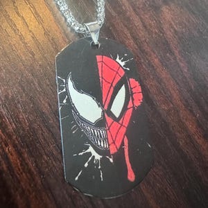 May include: A silver-colored chain necklace with a dog tag pendant featuring a split design. One side displays Venom's face in white and black, the other side shows Spider-Man's face in red and black.