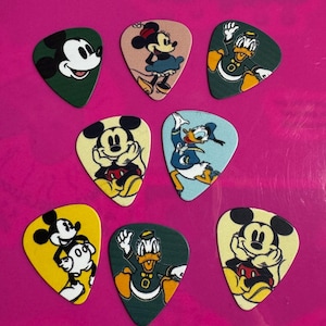 May include: Eight guitar plectrums featuring Disney characters. Mickey Mouse, Minnie Mouse, and Donald Duck are depicted in various poses. The picks have different coloured backgrounds, including green, yellow, and blue. Each pick is a different design.