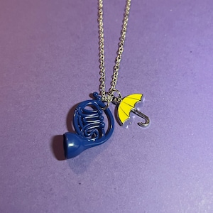 May include: A silver-colored necklace with two charms: a blue French horn and a yellow umbrella. The charms are suspended from a delicate chain against a purple background. The necklace is a piece of jewelry.