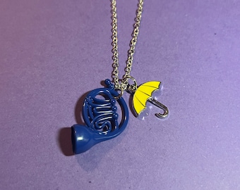 May include: A silver-colored necklace with two charms: a blue French horn and a yellow umbrella. The charms are suspended from a delicate chain against a purple background. The necklace is a piece of jewelry.