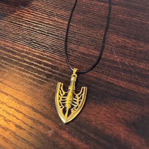 May include: A gold-coloured pendant necklace with a stylized arrow design. The pendant hangs from a black cord. The arrow has a shield-like shape with intricate detailing and a central, cylindrical element. The necklace is displayed on a wooden surface.