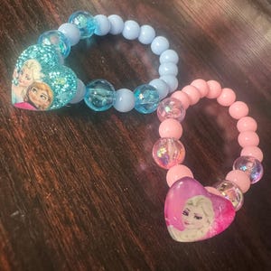Könnte beinhalten: Zwei Armbänder mit Perlen. Eines ist hellblau mit einem herzförmigen Anhänger mit zwei Zeichentrickfiguren. Das andere ist rosa mit einem herzförmigen Anhänger mit einer Zeichentrickfigur. Beide Armbänder haben klare, irisierende Perlen.