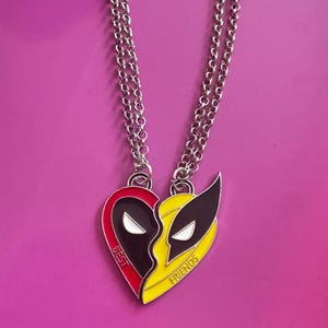 Könnte beinhalten: Eine silberne Kettenkette mit einem herzförmigen Anhänger. Der Anhänger ist in zwei Hälften geteilt, eine Seite zeigt ein rotes und schwarzes Deadpool-Design und die andere Seite ein gelbes und schwarzes Wolverine-Design. Der Anhänger trägt die Aufschrift "Beste Freunde" unten.