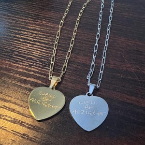 May include: Two heart-shaped pendant necklaces. One is gold-toned, the other silver-toned. Each pendant has the handwritten text "WE'LL BE ALRIGHT" engraved on the front. The necklaces have chain links.