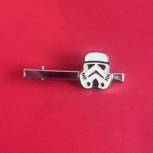 May include: A silver-colored tie clip featuring a white and black Stormtrooper helmet design. The clip is rectangular with a hinged clasp and is set against a red background. A Star Wars themed accessory.
