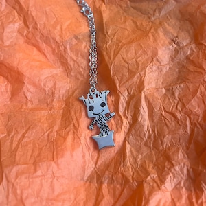 May include: Silver necklace featuring a charm of a cartoon character, Groot from Guardians of the Galaxy, standing in a pot. The charm is attached to a silver chain.