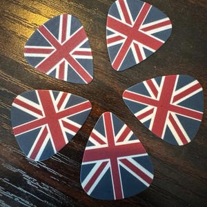 Könnte beinhalten: Fünf Gitarrenplektren mit Union Jack-Design. Jedes Plektrum zeigt die rot, weiß und blau Farben der britischen Flagge. Sie liegen auf einer dunklen Holzoberfläche.