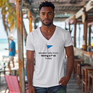 May include: White V-neck t-shirt with a blue flag graphic and the text "Marked Safe From Giving a F*ck Today". The shirt is made of a soft, comfortable material.