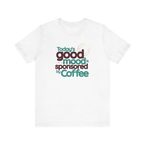 May include: White t-shirt featuring the slogan "Today's good mood is sponsored by Coffee" in teal, burgundy, and brown. The design includes a coffee cup ring. The shirt has a crew neck and short sleeves.
