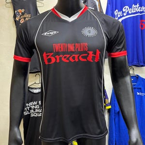 Pode incluir: Uma camisola preta de manga curta com acabamento vermelho nas mangas e gola em V. A frente apresenta o texto "TWENTY ONE PILOTS" em vermelho, "BreacH" numa fonte vermelha estilizada, um logótipo "top" branco e um emblema circular.