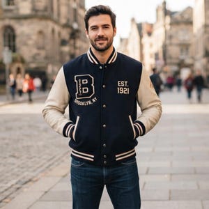 Può includere: Una giacca varsity blu navy con maniche color crema, caratterizzata da una grande lettera B con il testo Brooklyn, NY ed Est. 1921 ricamato sul davanti. La giacca presenta una chiusura con bottoni a pressione e bordi a coste a righe sul colletto.