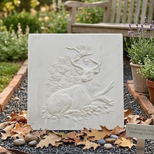 May include: A square off-white concrete stepping stone featuring a raised relief carving of a deer resting in front of a pine tree. The surface displays a detailed texture of the animal's fur, antlers, and the surrounding pine needles and cones.