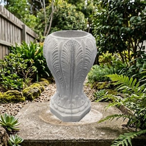 May include: A light gray concrete planter with a textured surface featuring vertical leaf-like carvings and a wide, circular opening at the top. The base of the planter is hexagonal, and the exterior displays a weathered, stone-like finish.