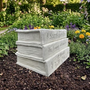 May include: A light gray concrete garden planter shaped like a stack of three books. The spines feature engraved text reading A Secret Garden, The Language of Flowers, and A Heart Full of Words, with textured page edges on the sides.