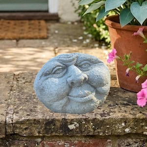May include: A gray stone garden ornament shaped like a round face with prominent cheeks, a wide nose, and a subtle smile. The sculpture features detailed carved eyes and a furrowed brow, crafted from a textured material resembling weathered concrete.