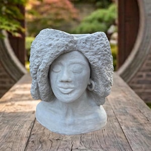 May include: A light gray concrete planter shaped like a human head with closed eyes and a textured, curly hairstyle. The sculpture features a smooth facial surface, defined lips, and a small hoop earring on the right side, with an open top.