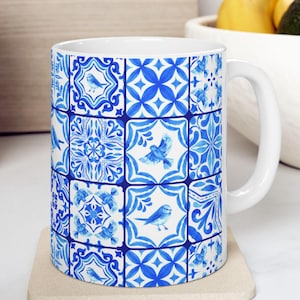 May include: White ceramic mug with a blue and white patterned design featuring a repeating tile pattern with birds. The mug has a handle and is sitting on a beige coaster.