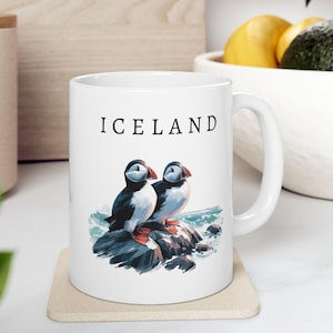 May include: White ceramic mug with the word "ICELAND" printed in black. The mug features a colorful illustration of two puffins perched on a rock, with blue water in the background. The mug sits on a beige coaster.